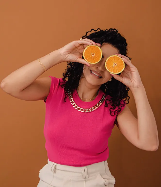 Amanda Lima sorrindo, segurando duas metades de laranja diante dos olhos, com blusa rosa e fundo caramelo
