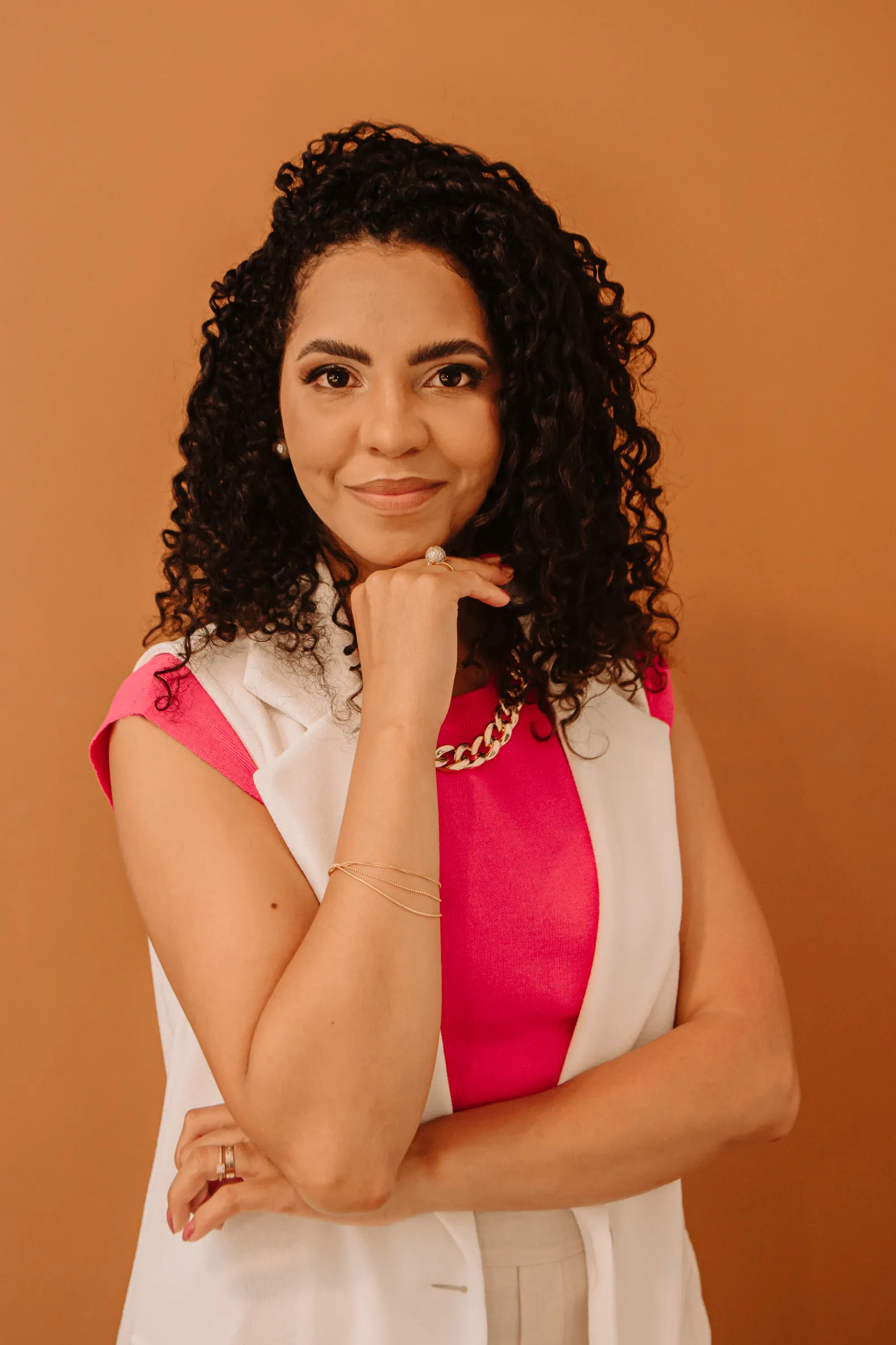 Amanda Lima, nutricionista, sorrindo com a mão no queixo, de colete branco e blusa rosa, diante de um fundo caramelo