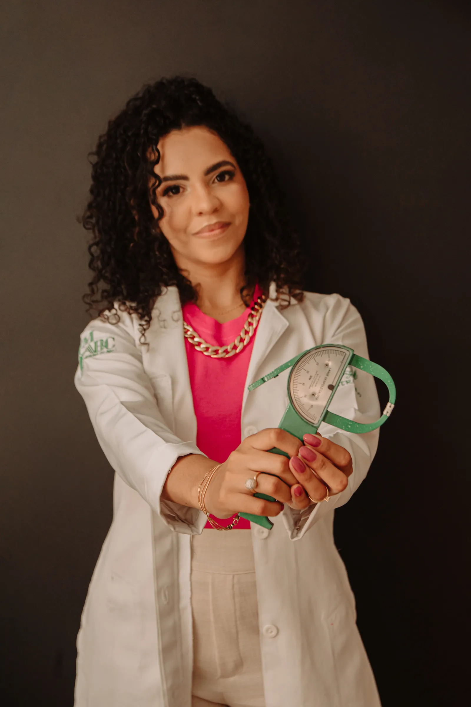 Amanda Lima de jaleco branco e blusa rosa, estendendo um adipômetro verde à frente, diante de um fundo escuro