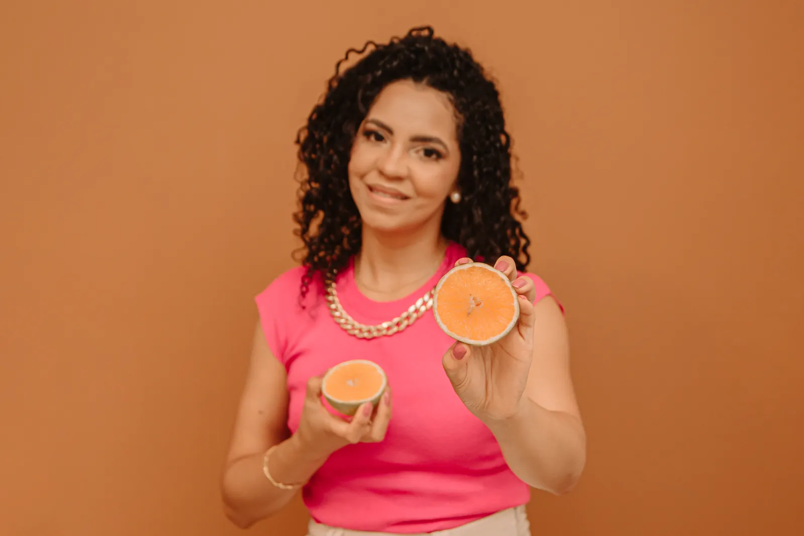 Amanda Lima sorrindo, segurando duas metades de laranja, de blusa rosa e fundo caramelo