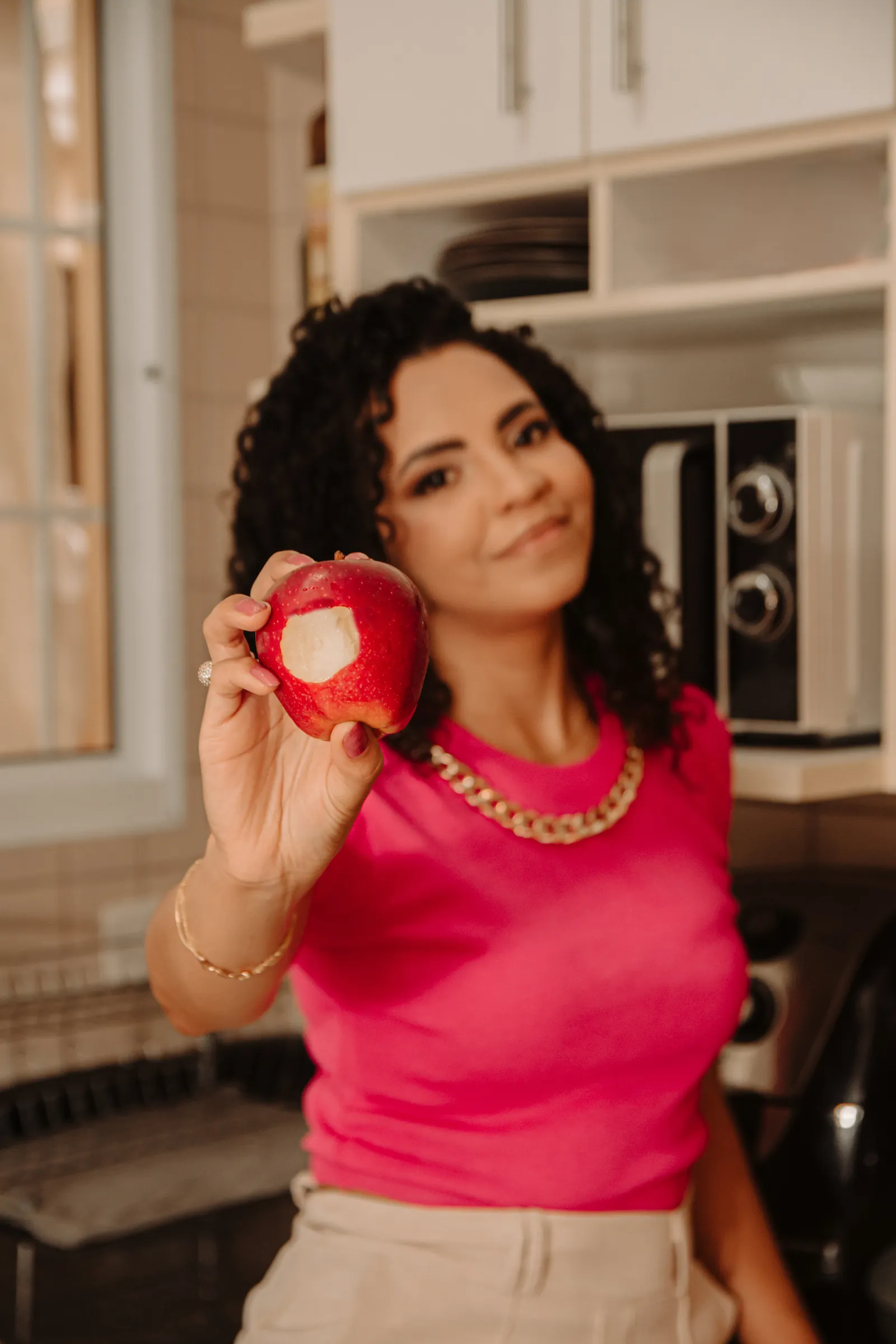 Amanda Lima de blusa rosa segurando uma maçã mordida em uma cozinha