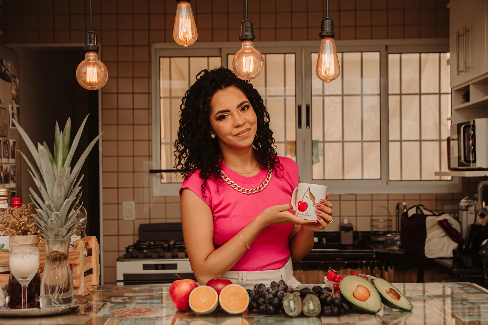 Amanda Lima em uma cozinha, segurando uma caneca, com frutas sobre a bancada à frente