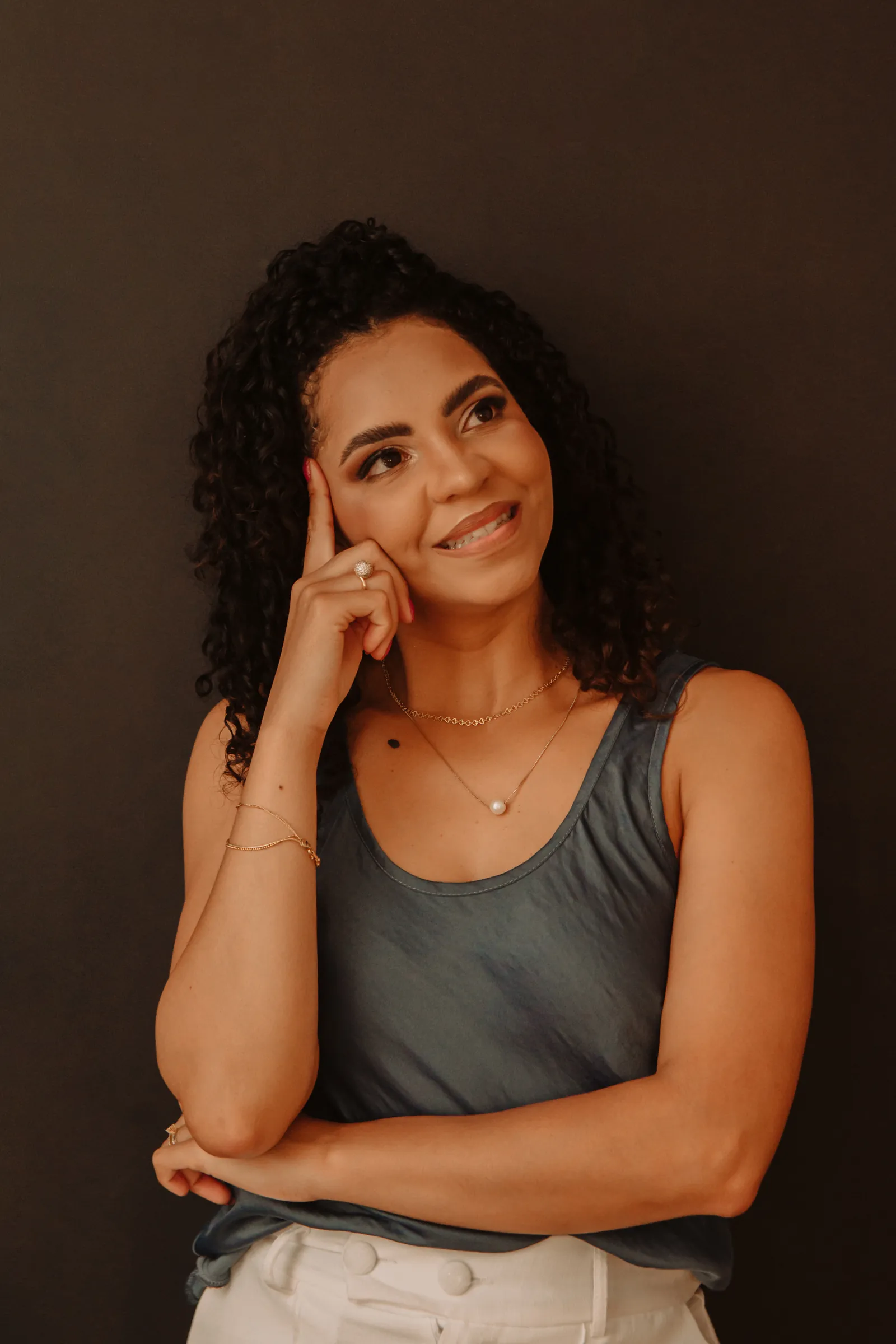 Amanda Lima sorrindo, pensativa, com a mão na têmpora, de regata cinza diante de um fundo escuro