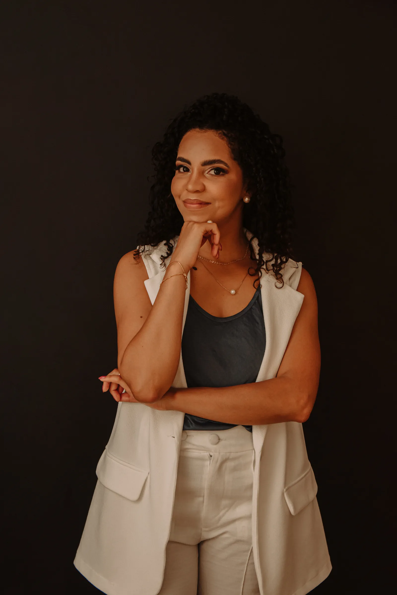 Amanda Lima de colete branco, sorrindo com a mão no queixo, diante de um fundo escuro
